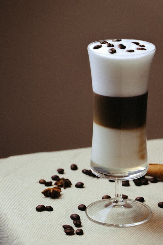 Stylish latte macchiato with cream foam, elegantly presented in a tall glass surrounded by scattered coffee beans.