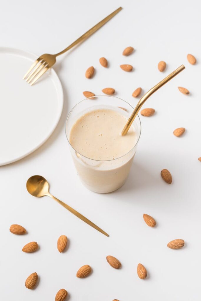 A creamy almond smoothie on a white background, styled with golden utensils and scattered almonds.