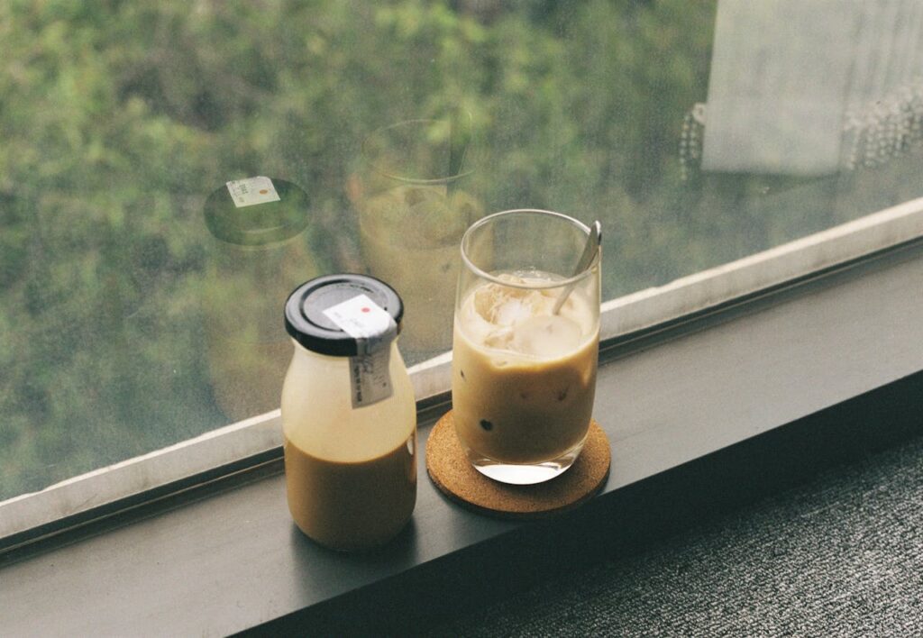 A refreshing iced beverage and milk bottle placed on a windowsill with a view of greenery.